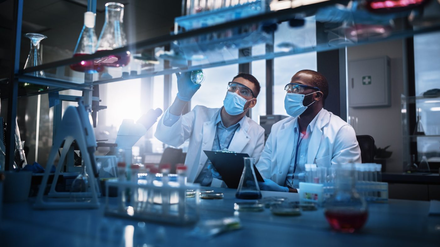 Modern Medical Research Laboratory: Two Scientists Wearing Face Masks use Microscope, Analyse Sample in Petri Dish, Discuss Innovative Technology. Advanced Scientific Lab for Medicine, Biotechnology