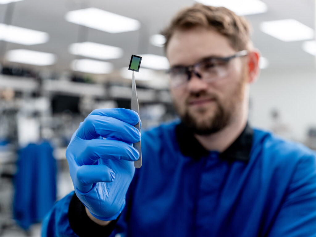 Male scientists holding a chip technology the size of a nickle on tweezers with blue gloves.