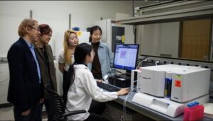 (Standing, from left) Rong Tong, associate professor in chemical engineering; Wenjun "Rebecca" Cai, associate professor in materials science and engineering; Eungyo Jang; and Ziyu Huo gather around Liqian Niu (seated), who is working with a Luminex 200 machine used to analyze tumor cytokine levels.